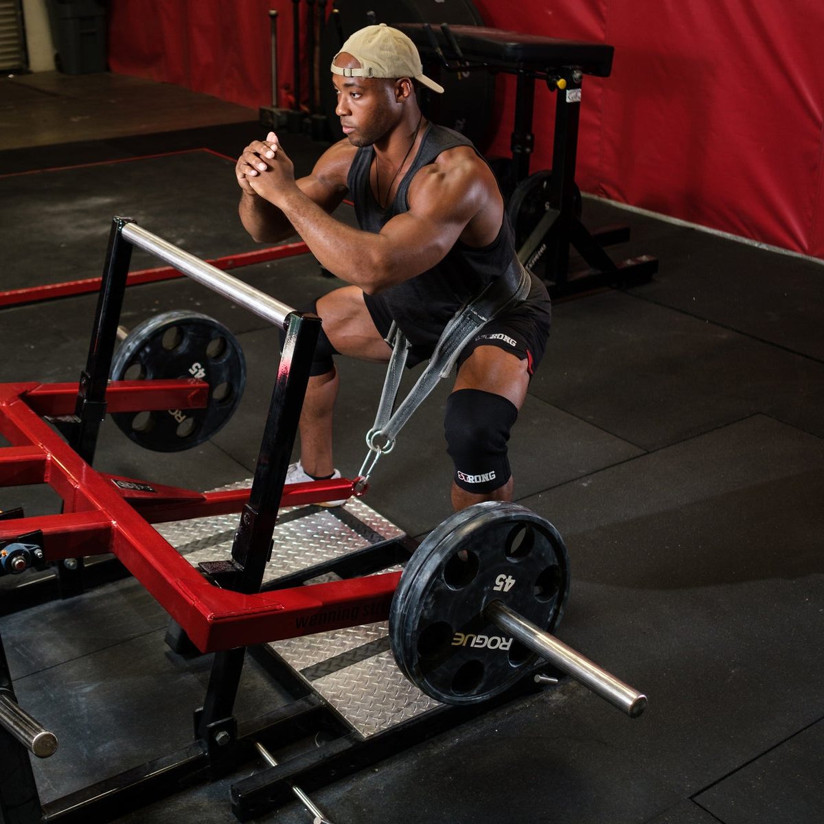 A lifter wearing black STrong knee sleeves at the bottom of a squat on a belt squat machine, in a gym with red and black equipment.